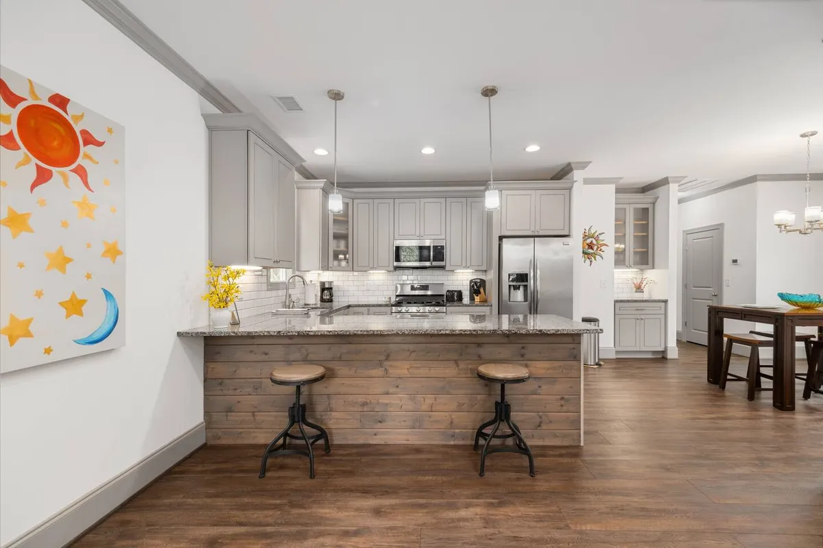 A bright, welcoming kitchen scene featuring a central island with stools, modern appliances, and tasteful artwork on the wall. The layout is open, with visible space for both cooking and socializing.