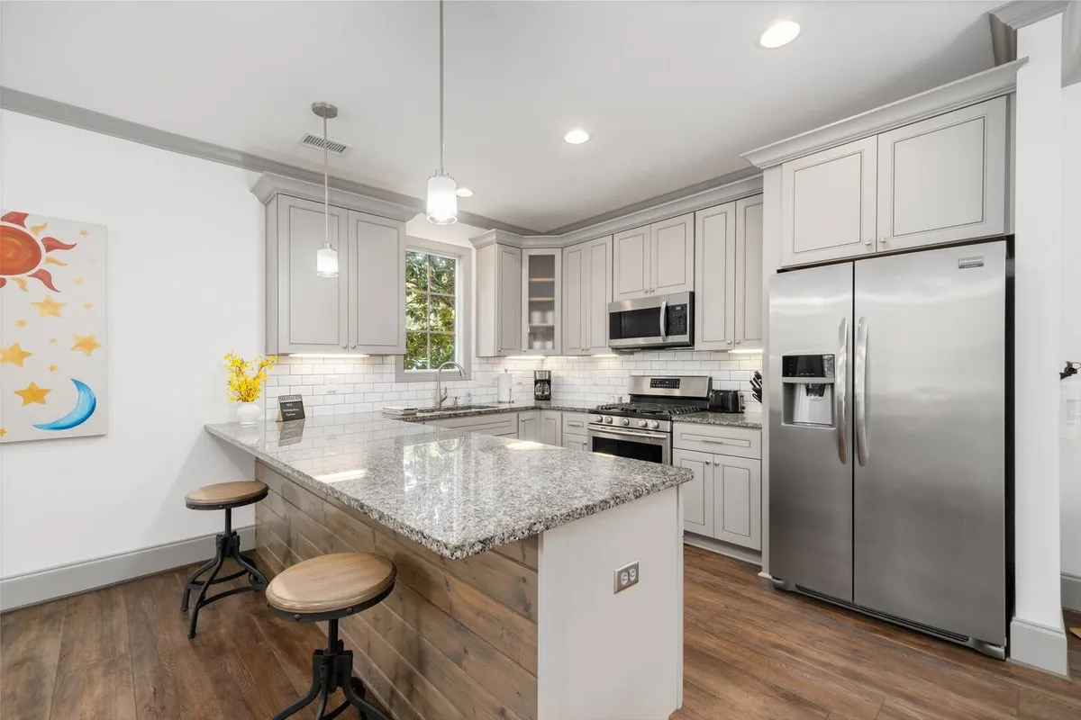 A modern kitchen with granite countertops, stainless steel appliances, and cheerful natural light streaming through a window. Colorful wall art adds a personalized touch, and the setup suggests a space built for entertaining and family meals.