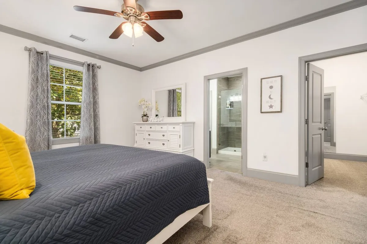 A serene bedroom featuring a plush bed with a navy quilt and a yellow accent pillow, complemented by a dresser with a mirror. A large window with curtains allows for adjustable natural light, reinforcing the sense of private comfort and relaxation.