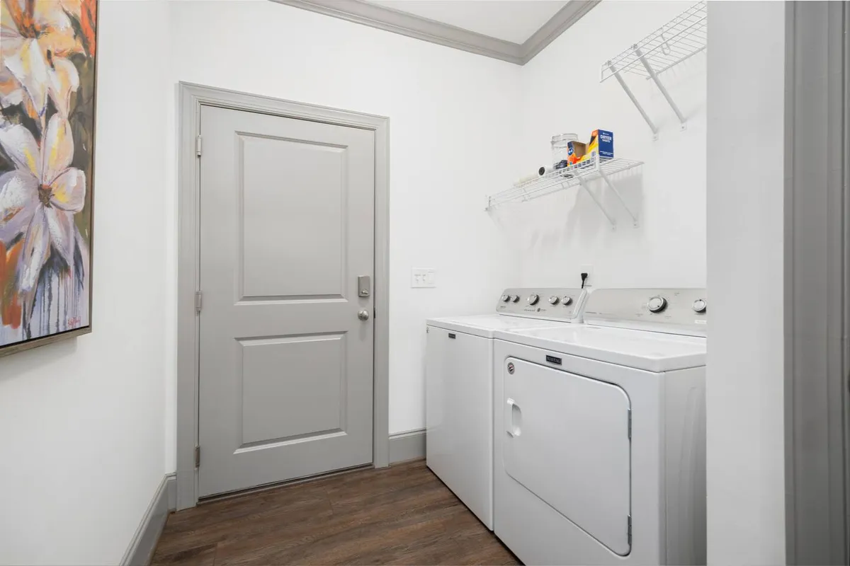 A practical laundry area featuring a washing machine, dryer, shelving with supplies, and a door that hints at movement between indoor and outdoor activities. The scene is functional and efficient, suggesting ease and comfort after an active day.