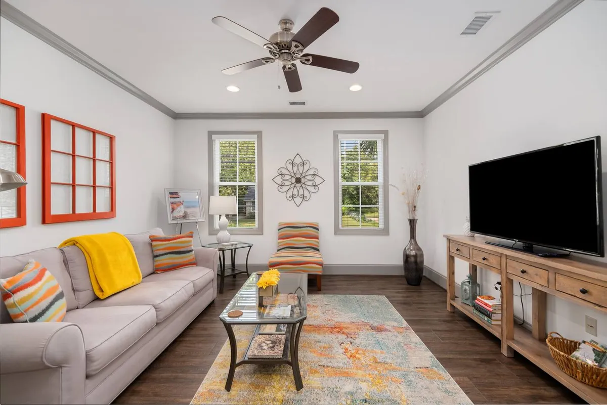 A vibrant living area featuring a modern sofa adorned with a yellow throw and colorful cushions, a sleek glass coffee table with decorative items, a TV stand with decor and lush plants, and bold, colorful artwork on the walls. Sunlight streams through the windows, enhancing the contemporary atmosphere and highlighting the open layout.