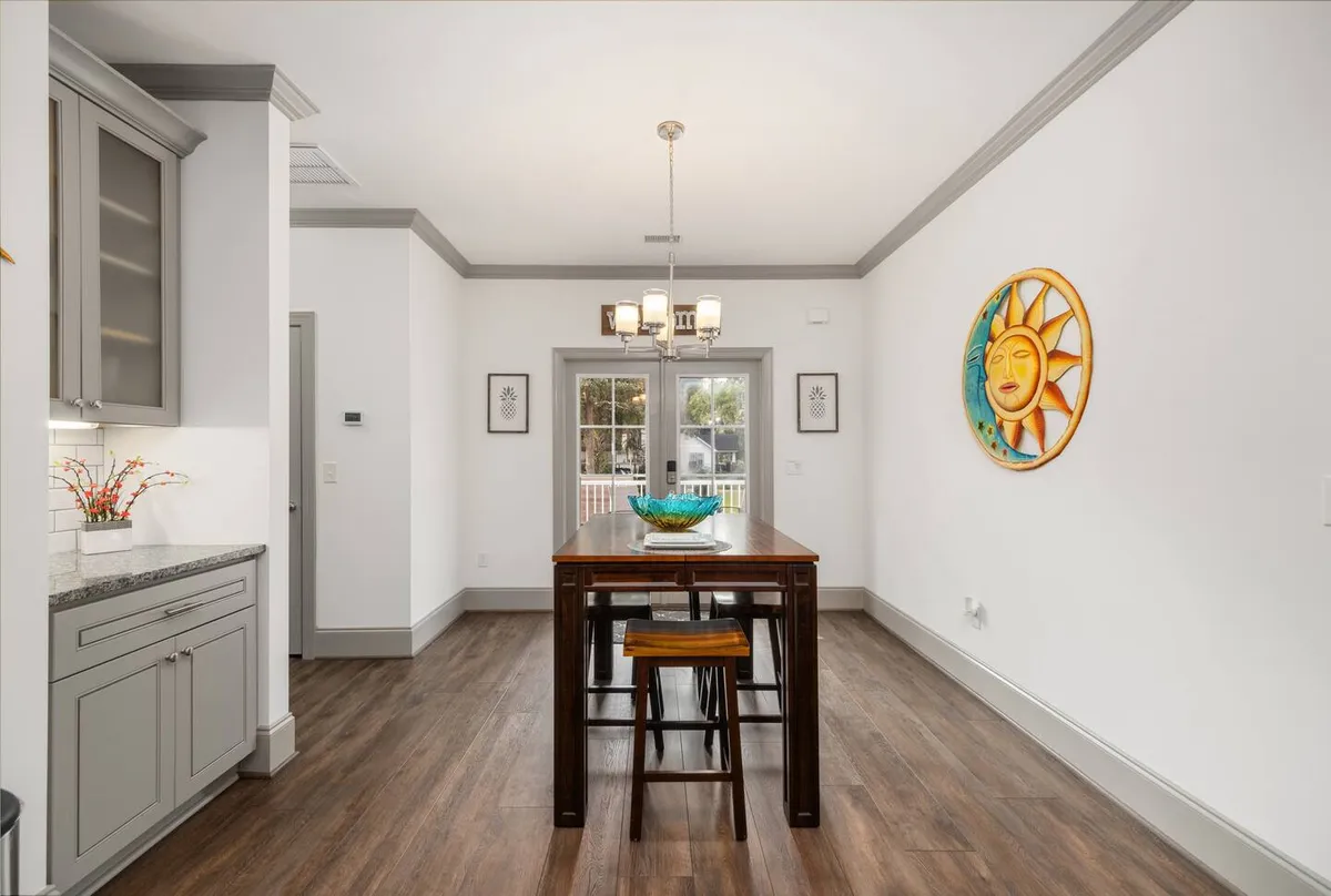 A cheerful dining area with a central dining table, colorful wall art, an elegant chandelier, and a glimpse of kitchen cabinets in the background. The space is bathed in natural light, fostering an inviting and festive mood suitable for shared meals or morning coffee.